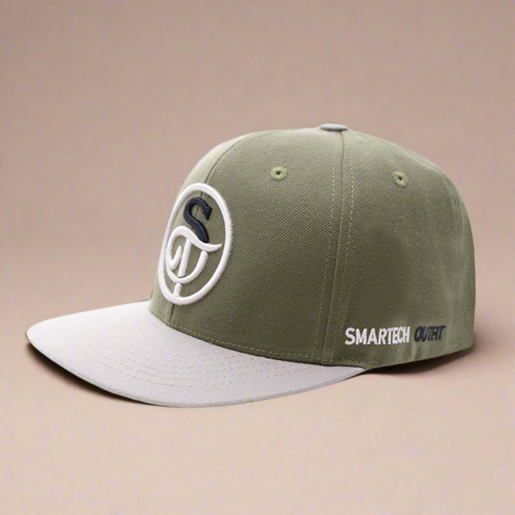 An olive green and beige snapback cap, featuring a curved white insignia with a stylized "S" on the front. Made from high-quality acrylic, the brim is beige and the side of the cap has "SMARTECH OUTFIT" in white and black letters, emphasizing brand quality.